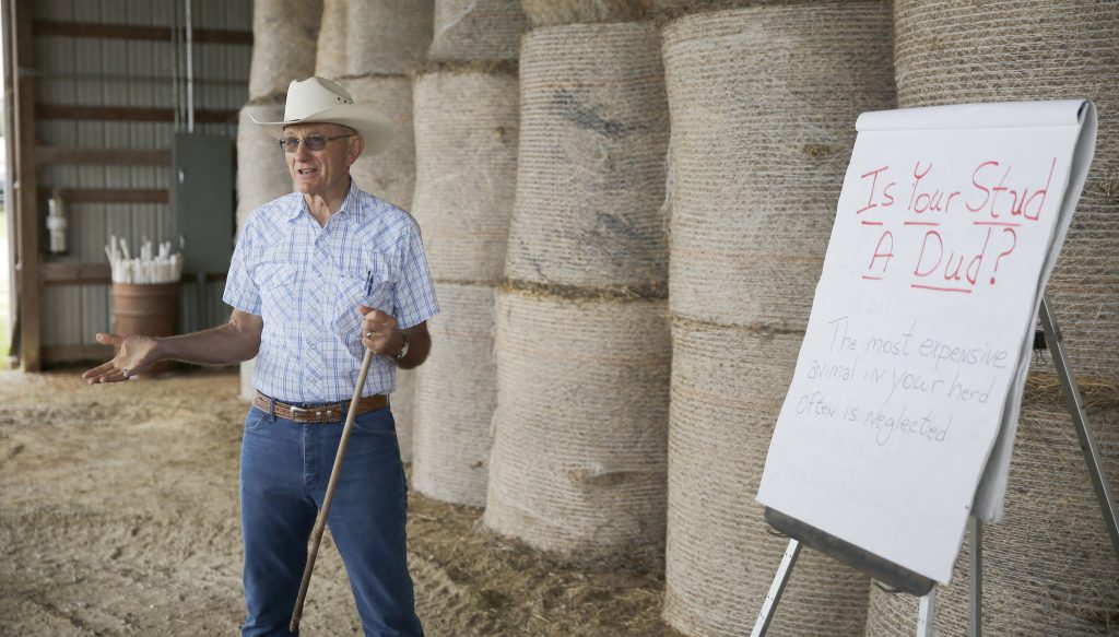 An older white man in a white cowboy hat, short-sleeved button-up shirt, and blue jeans speaks next to a sign that reads, "Is your stud a dud?"